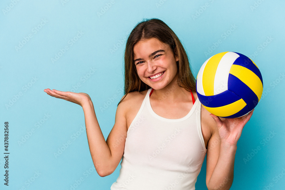 Young caucasian woman playing volleyball isolated on blue background showing a copy space on a palm and holding another hand on waist.