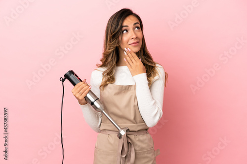 Young woman using hand blender over isolated pink background looking up while smiling