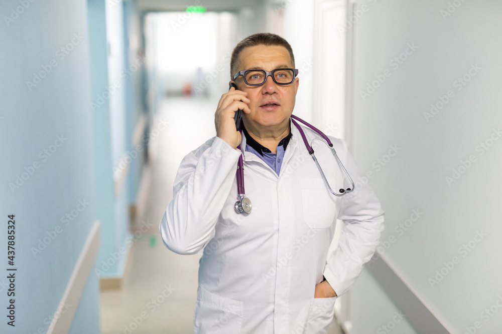 Shot of male doctor talking with somebody on his mobile phone while standing on the clinic foyer. Shot of male doctor talking with somebody on his mobile phone while standing on the clinic foyer.