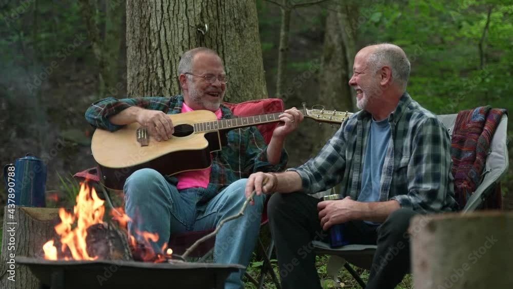 Two mature gay men playing guitar in front of a campfire and laughing while camping in a forest.