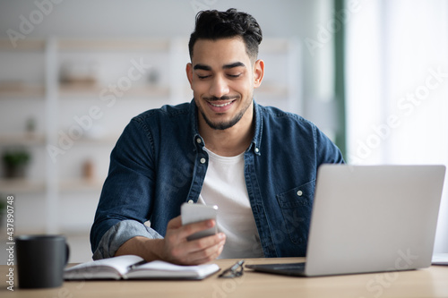 Tela Cheerful arab guy using smartphone while sitting at workdesk