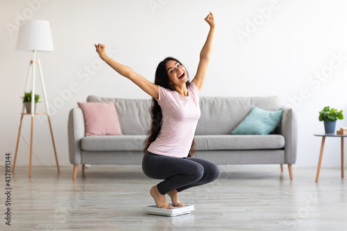 Excited Indian woman sitting on scales, raising hands up, happy with result of her weight loss program at home