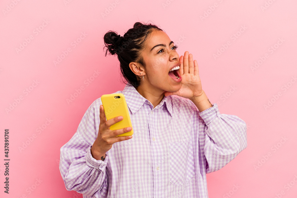 Young latin woman holding a mobile phone isolated on pink background shouting and holding palm near opened mouth. Young latin woman holding a mobile phone isolated on pink background shouting and holding palm near opened mouth.