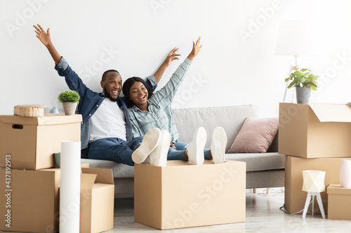 Housewarming Party. Joyful Black Couple Sitting Among Cardboard Boxes And Raising Hands