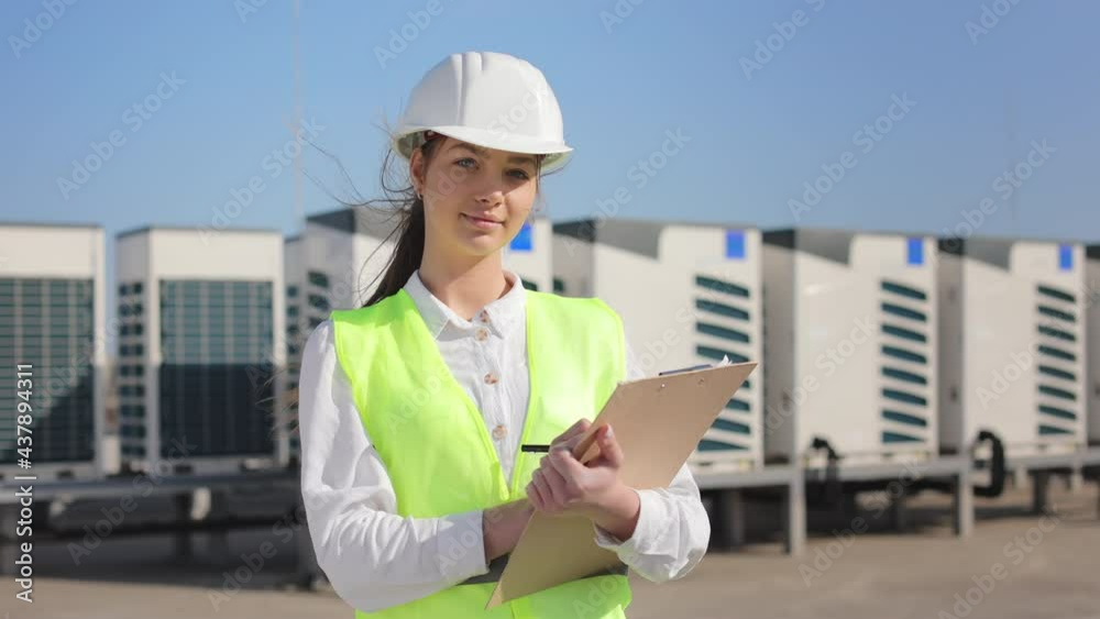 Portrait of a happy woman engineer is making entries in documents and looking at the camera. She's on the roof of a business center. She is wearing work clothes and a hard hat. Air conditioning Portrait of a happy woman engineer is making entries in documents and looking at the camera. She's on the roof of a business center. She is wearing work clothes and a hard hat. Air conditioning
