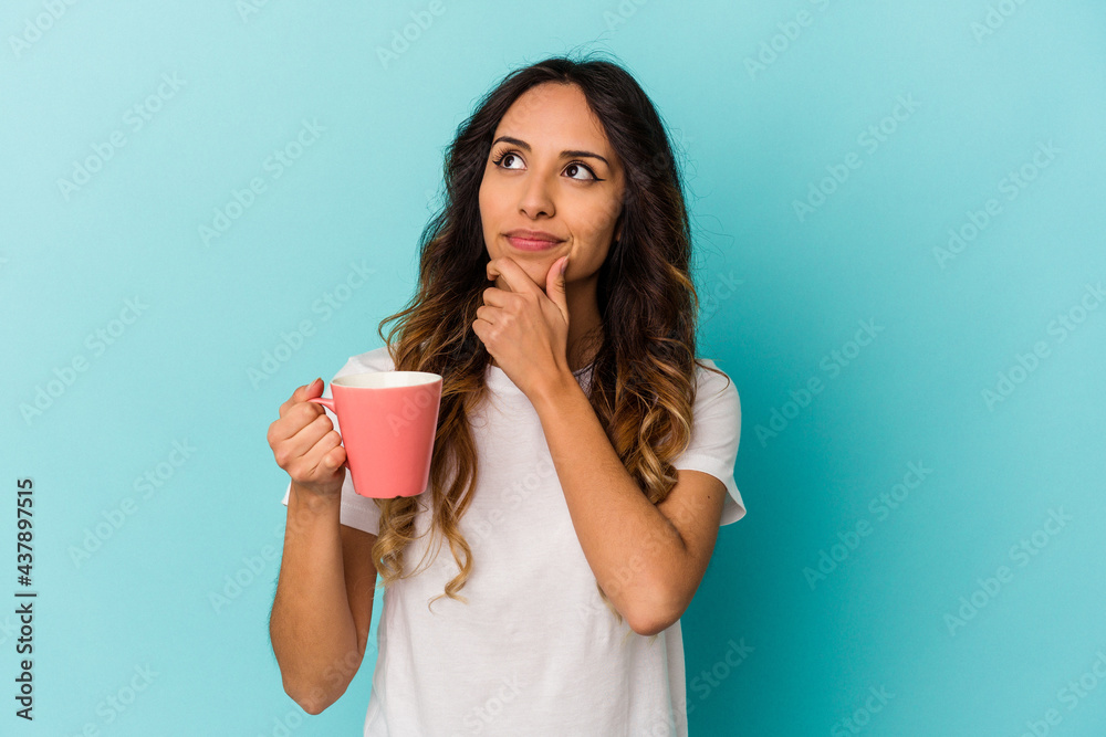 Young mexican woman holding a mug isolated on blue background looking sideways with doubtful and skeptical expression. Young mexican woman holding a mug isolated on blue background looking sideways with doubtful and skeptical expression.