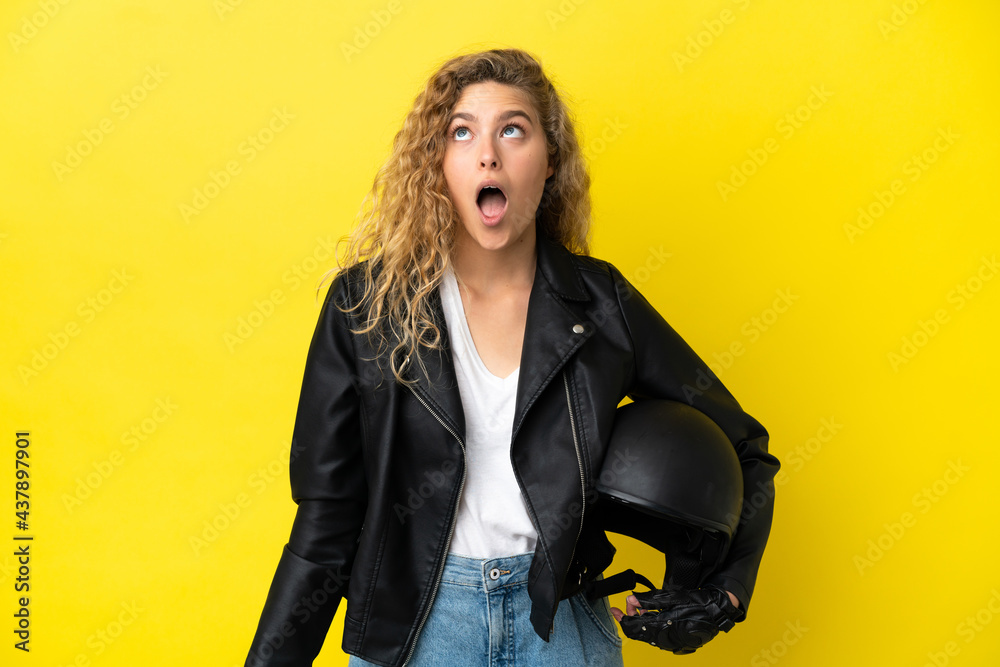 Young blonde woman with a motorcycle helmet isolated on yellow background looking up and with surprised expression Young blonde woman with a motorcycle helmet isolated on yellow background looking up and with surprised expression