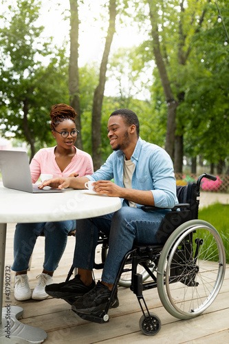 Happy disabled black man and his female coworker using laptop together at table in outdoor cafe, copy space © Prostock-studio
