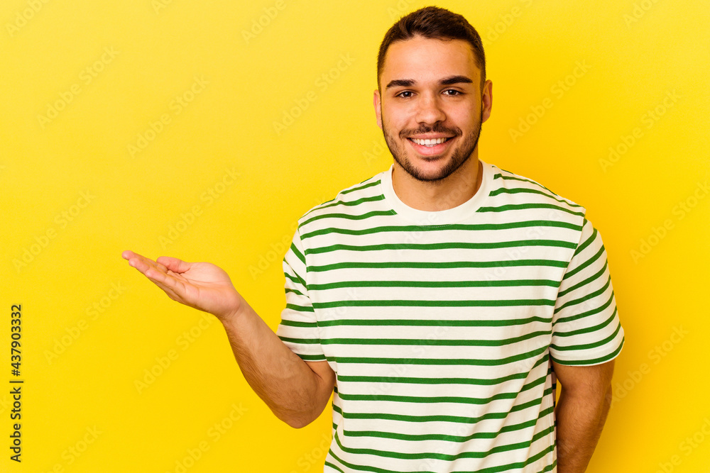 Young caucasian man isolated on yellow background showing a copy space on a palm and holding another hand on waist. Young caucasian man isolated on yellow background showing a copy space on a palm and holding another hand on waist.
