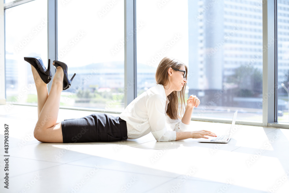 Obraz premium Beautiful female specialist with laptop computer laying on the floor in modern office and smiling charmingly. Working on design, data analysis, plan strategy. Business people concept