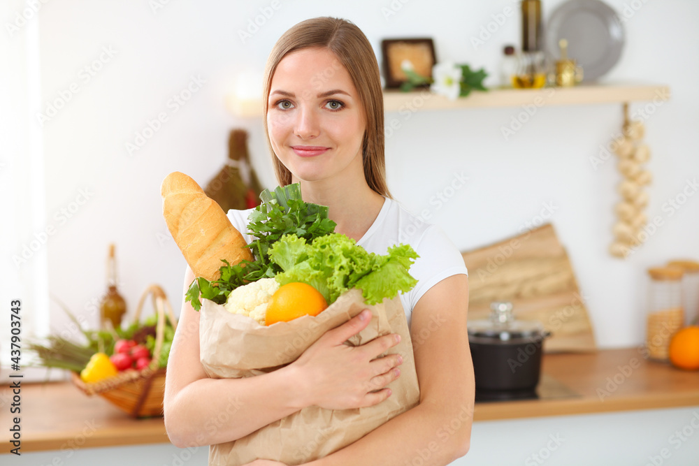 An attractive young woman holding the paper bag full of vegetables while standing and smiling in sunny kitchen. Cooking concept