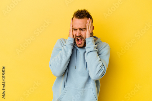 Young tattooed caucasian man isolated on yellow background covering ears with hands trying not to hear too loud sound.