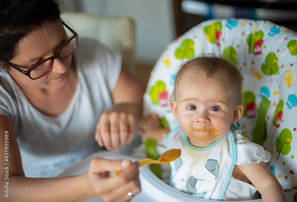 mom giving homogenized food to her daughter on high chair. Messy feeding mom giving homogenized food to her daughter on high chair. Messy feeding