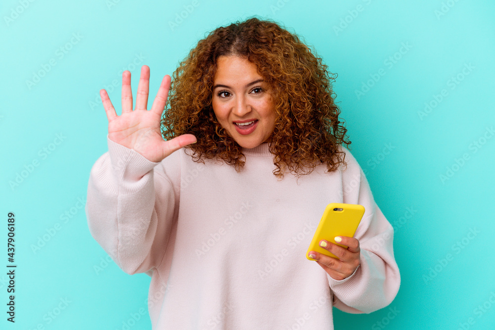 Young latin woman holding mobile phone isolated on blue background smiling cheerful showing number five with fingers.