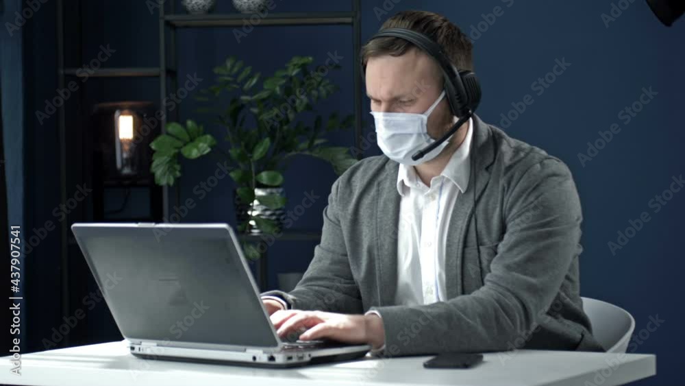 Office work during the coronavirus epidemic. Middle-aged businessman in headphones works with a laptop. Man has a protective mask on his face.