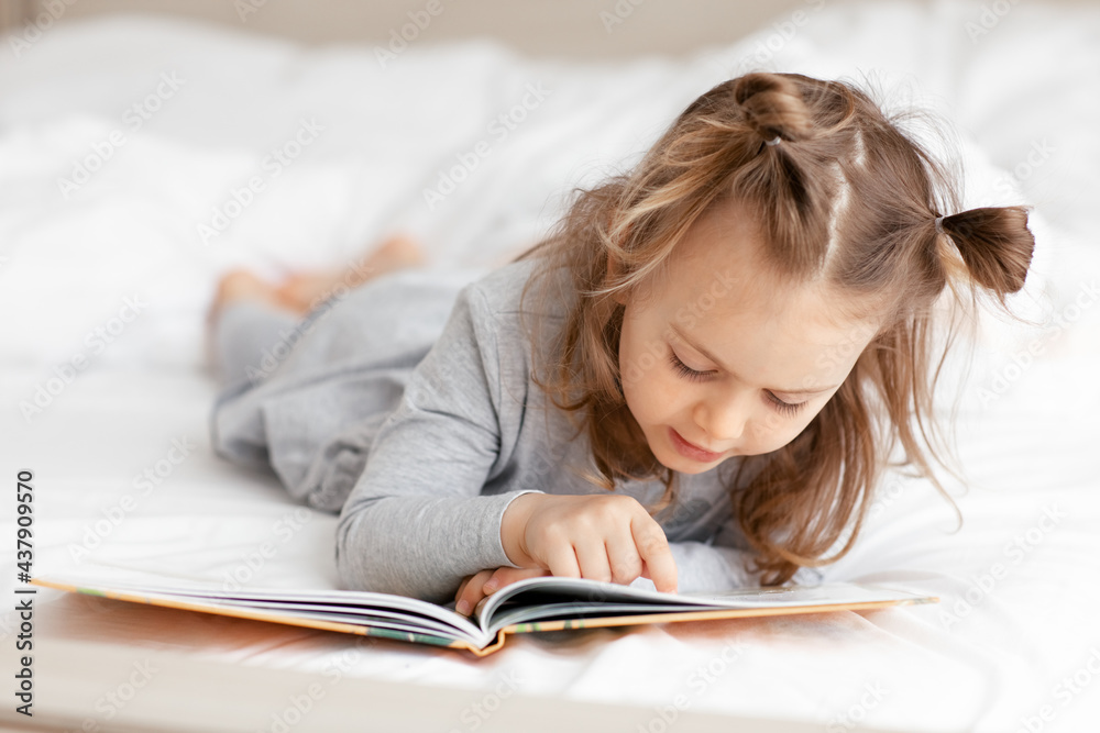 Focused child girl reading a book in bed. Concentrated kid girl looks into a book and moves her finger across the text.International literacy day. Focused child girl reading a book in bed. Concentrated kid girl looks into a book and moves her finger across the text.International literacy day.