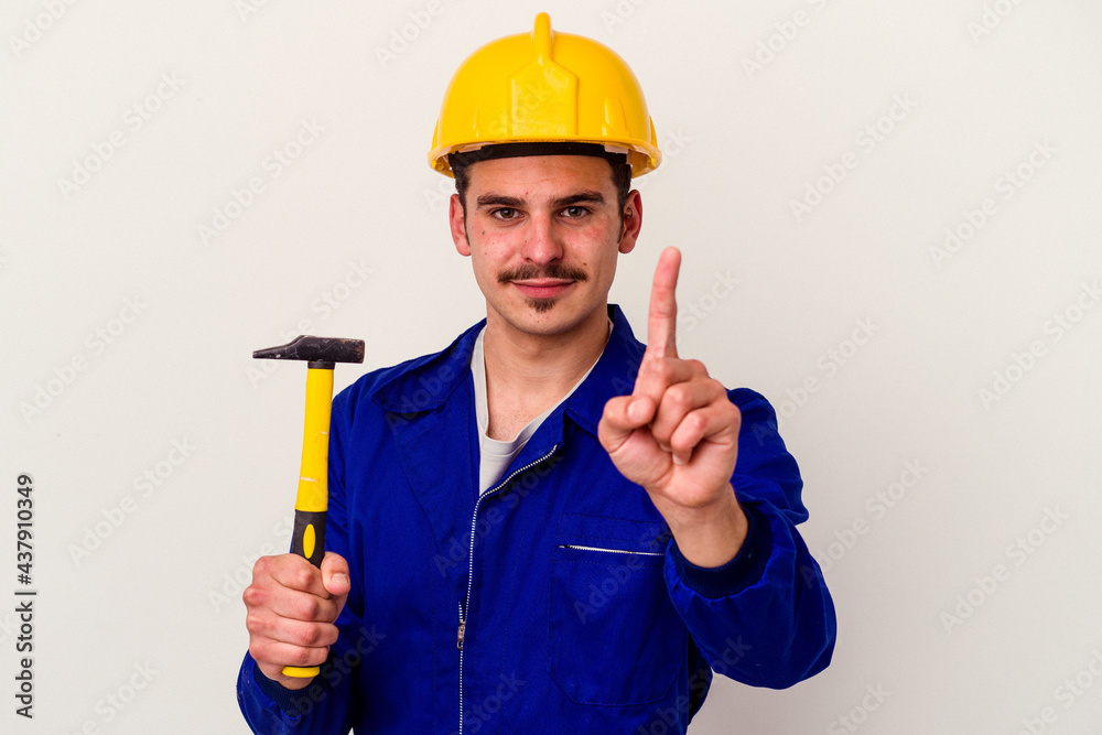 Young caucasian worker man holding a hammer isolated on white background showing number one with finger. Young caucasian worker man holding a hammer isolated on white background showing number one with finger.