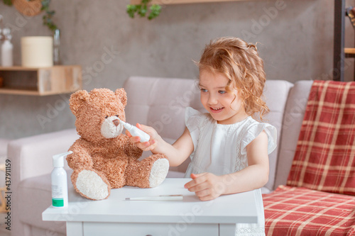a happy sweet little girl at home on the couch smiling, playing with a teddy bear in the doctor treats, puts on a thermometer, drips her nose