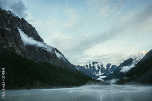 Scenic alpine landscape with snowy mountain peak in golden sunlight and mountain lake in fog under cloudy sky. Atmospheric highland scenery with sunlit mountain top and low clouds on rocks and slopes.