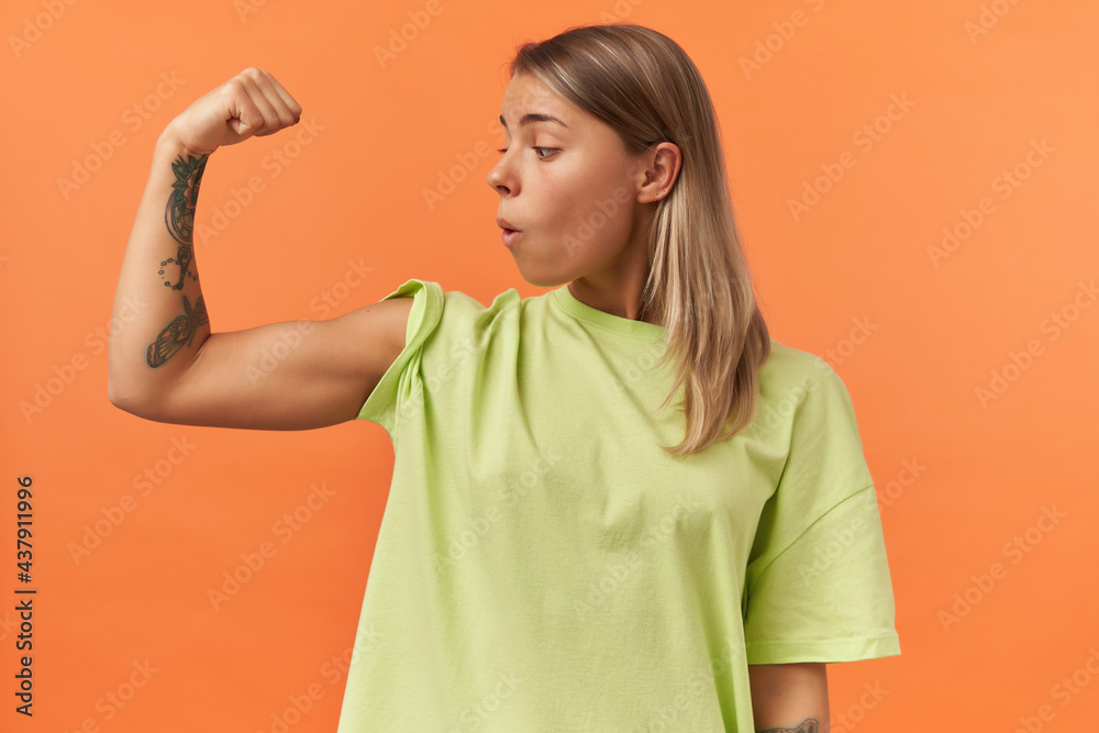 Surprised beautiful young woman in yellow tshirt showing biceps muscles and looking on it isolated over orange background Surprised beautiful young woman in yellow tshirt showing biceps muscles and looking on it isolated over orange background