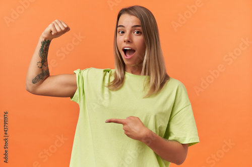 Amazed pretty young woman with opened mouth in yellow tshirt showing biceps muscles and pointing on it isolated over orange background