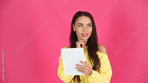 thoughtful young adult woman holding tablet with hand near face gesture isolated on pink