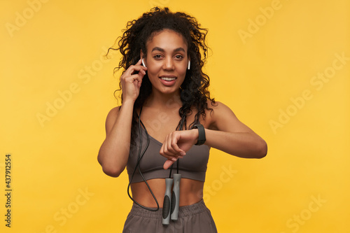 Smiling pretty african american young fitness woman with skipping rope on neck listening to music using wireless earphones and smart watch isolated over yellow background