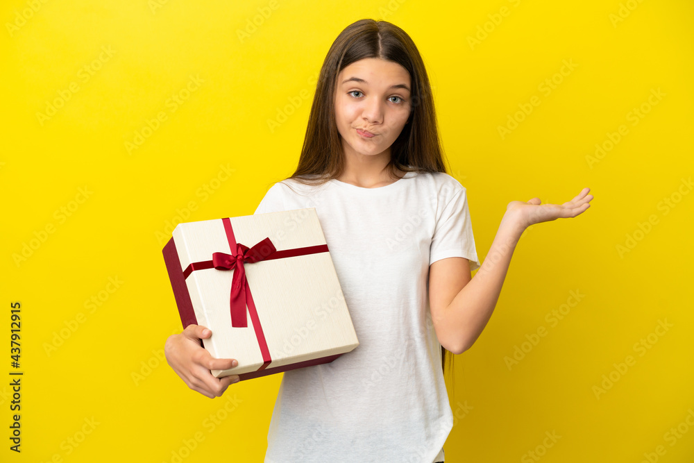 Little girl holding a gift over isolated yellow background having doubts while raising hands Little girl holding a gift over isolated yellow background having doubts while raising hands