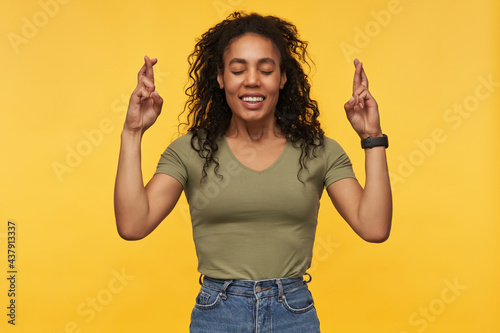 Happy attractive african american young woman in casual clothes keeps eyes closed and fingers crossed isolated over yellow background Making a wish