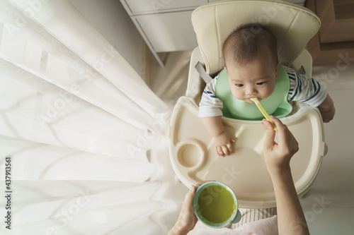 Fotografie Asian little cute baby boy sitting in high chair eating first food from spoon with his mother at home