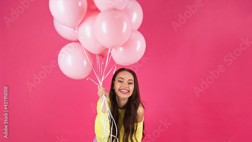 joyful young adult woman in yellow sweatshirt holding balloons isolated on pink