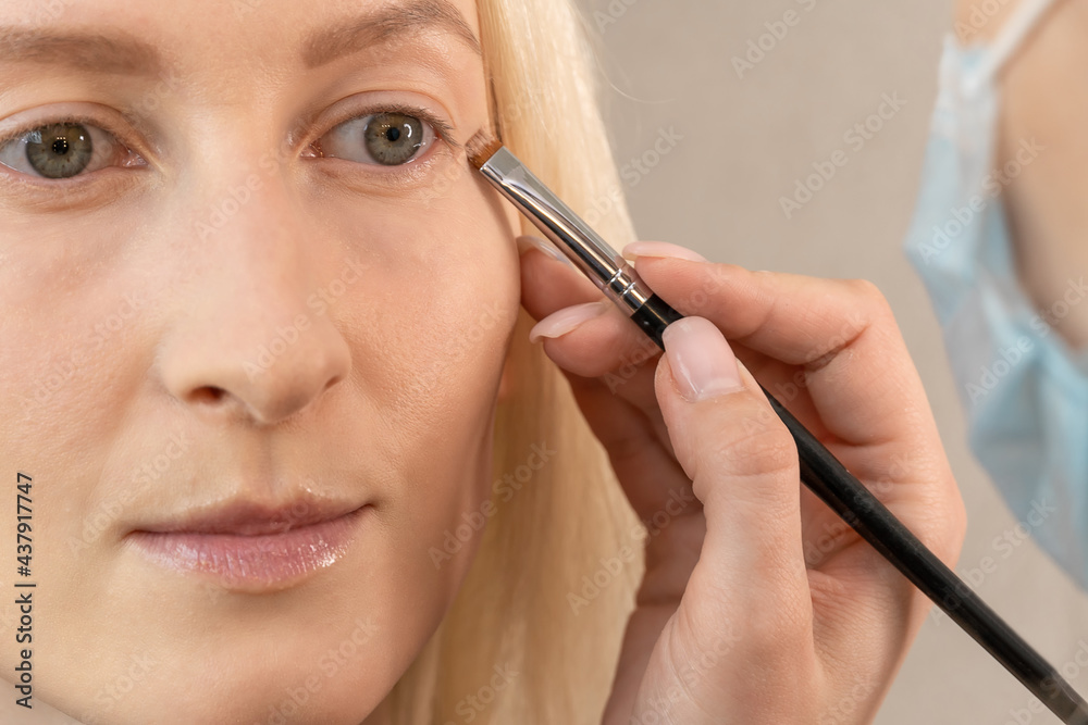 Make-up artist in a medical mask uses a brush to apply makeup base on the eyelid close-up Make-up artist in a medical mask uses a brush to apply makeup base on the eyelid close-up
