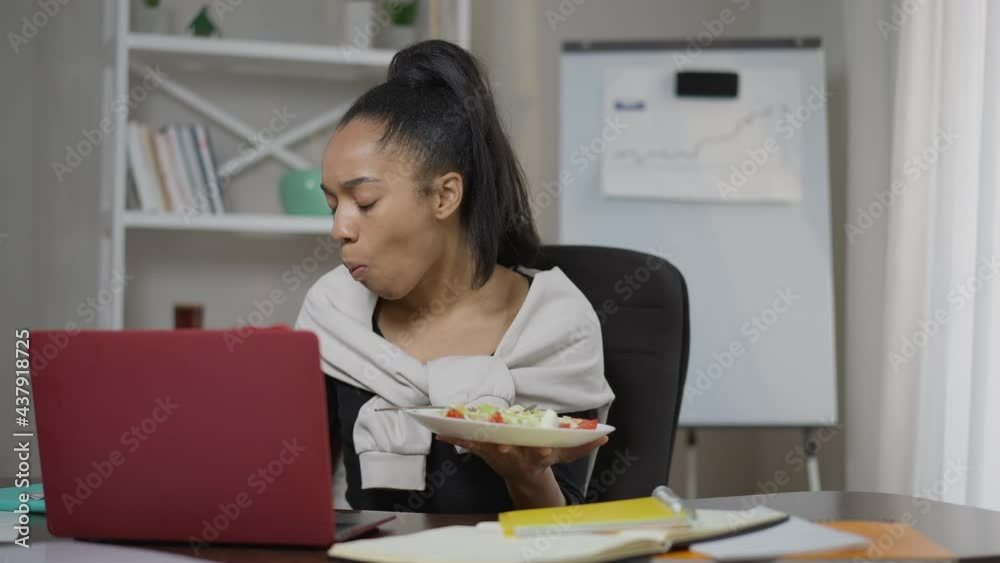 Front view portrait of busy young focused woman eating healthful veggie salad surfing Internet on laptop in home office. Concentrated smart African American lady overworking indoors. Multitasking