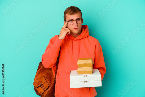 Young student caucasian man holding hamburgers and pizzas isolated on blue background