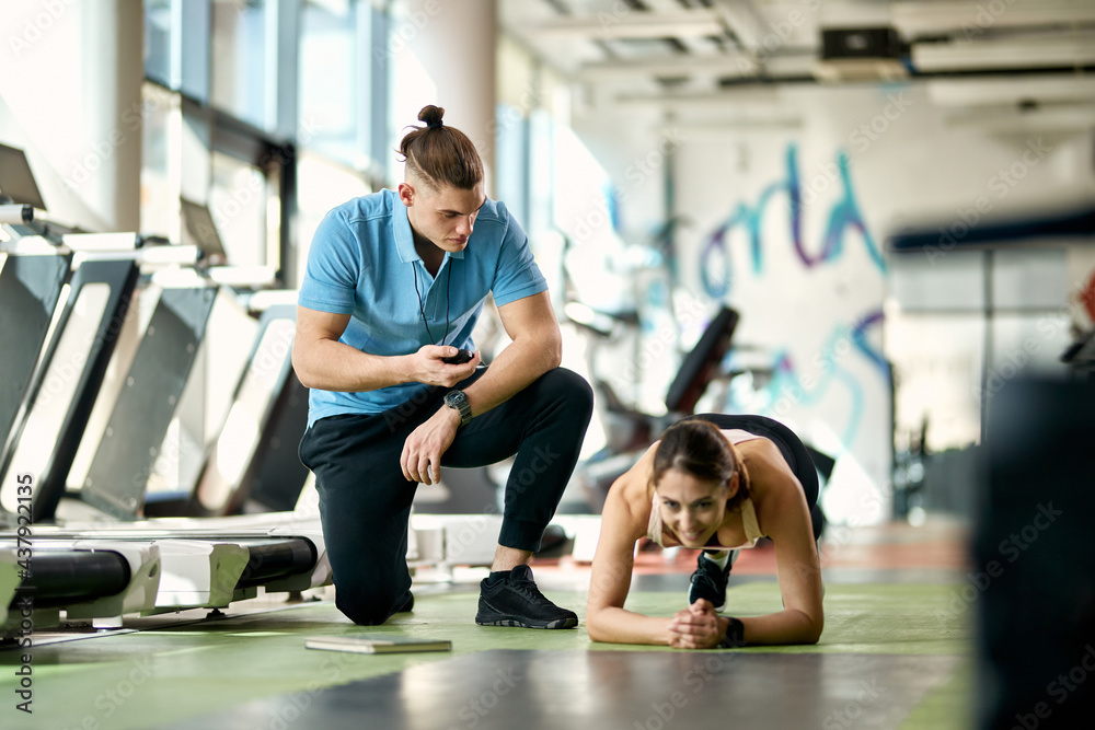 Athletic woman exercising planks while male coach is measuring time on stopwatch in a gym.