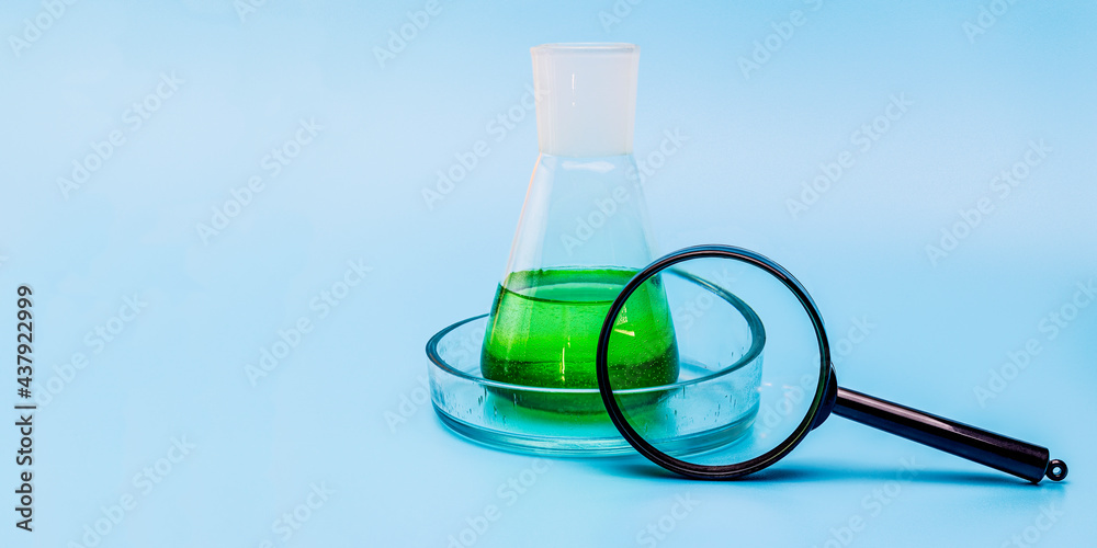 Glass laboratory flask in a cup with green liquid with a magnifying glass on a gentle blue medical background. The concept of medical and chemical experiments. Close-up. copy space. Glass laboratory flask in a cup with green liquid with a magnifying glass on a gentle blue medical background. The concept of medical and chemical experiments. Close-up. copy space.