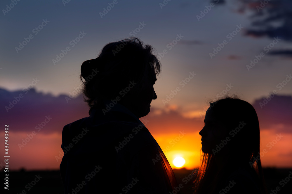 Silhouettes of an elderly woman and a girl on the background of the sunset. Granddaughter and grandmother look at each other. The concept of the family.