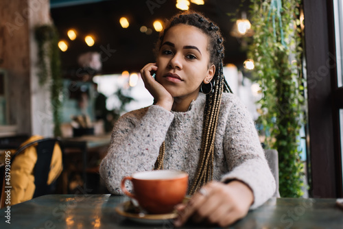 Portrait of attractive African American customer sitting at cafeteria table with tea cup and looking at camera,dark skinned hipster girl with Afro pigtails posing during leisure weekend in coffee shop