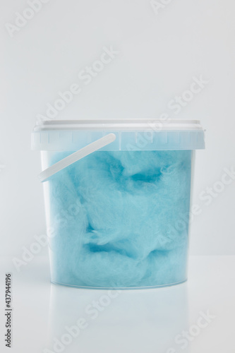 Blue sweet cotton candy in a transparent plastic bucket with a lid and a white handle on a white background.