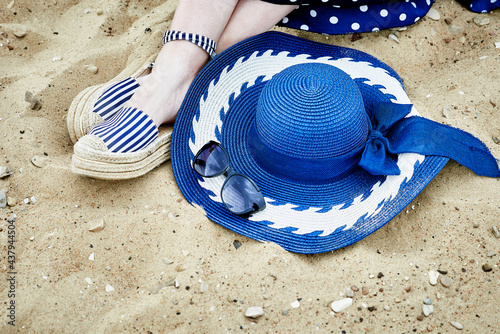 Women's feet in striped sandals, blue hat and sunglasses on the sand.