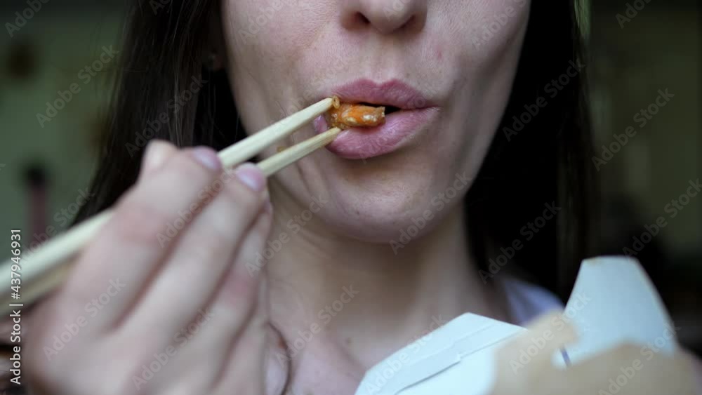 Close-up of a girl eating Asian food from a paper box with wooden chopsticks. Street food concept, quick bite Close-up of a girl eating Asian food from a paper box with wooden chopsticks. Street food concept, quick bite
