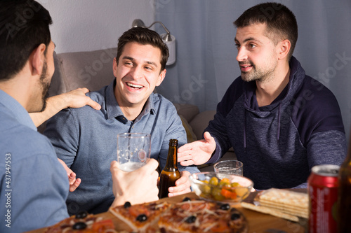 Three cheerful men talking and laughing while enjoying beer and pizza at home