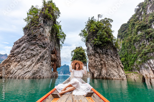 Young Female Tourist in Dress and Hat at Longtail Boat near Famous Three rocks with Limestone Cliffs at Cheow Lan Lake. Travel Woman Sitting on Boat in Khao Sok National Park in Thailand