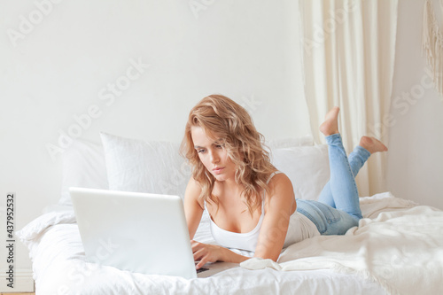 Female student sitting on bed of her apartment with laptop studying at home. Casual woman using laptop