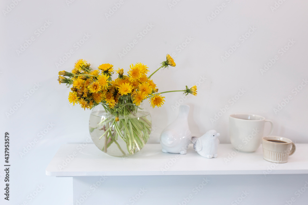 A bouquet of dandelions in a white fluted vase and two cups of different sizes with coffee on a white table. A bouquet of dandelions in a white fluted vase and two cups of different sizes with coffee on a white table.