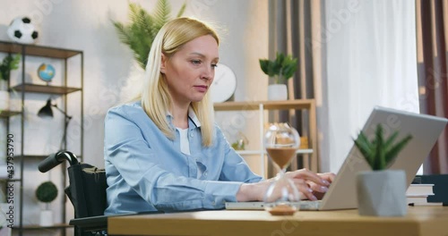 Attractive concentred modern professional businesswoman sitting in wheelchair after accident and working on computer at home in daytime photo