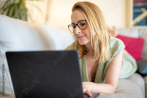 Attractive young woman working on laptop and smiling while lying on the sofa at home