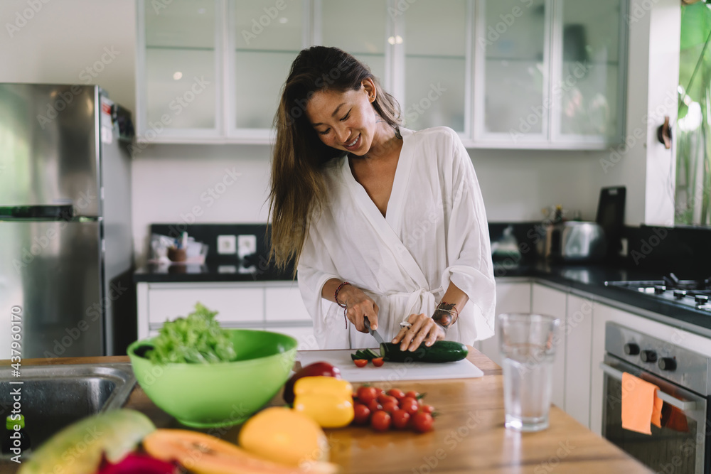 Stylish young ethnic female smiling while cutting vegetables in modern kitchen