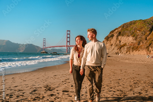 A young man and a woman take a romantic walk on the beach overlooking the Golden Gate Bridge at sunset in San Francisco