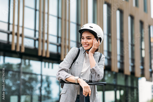 Positive woman leaning on handlebar of electric push scooter. Pretty female in helmet looking away while standing in the city. photo